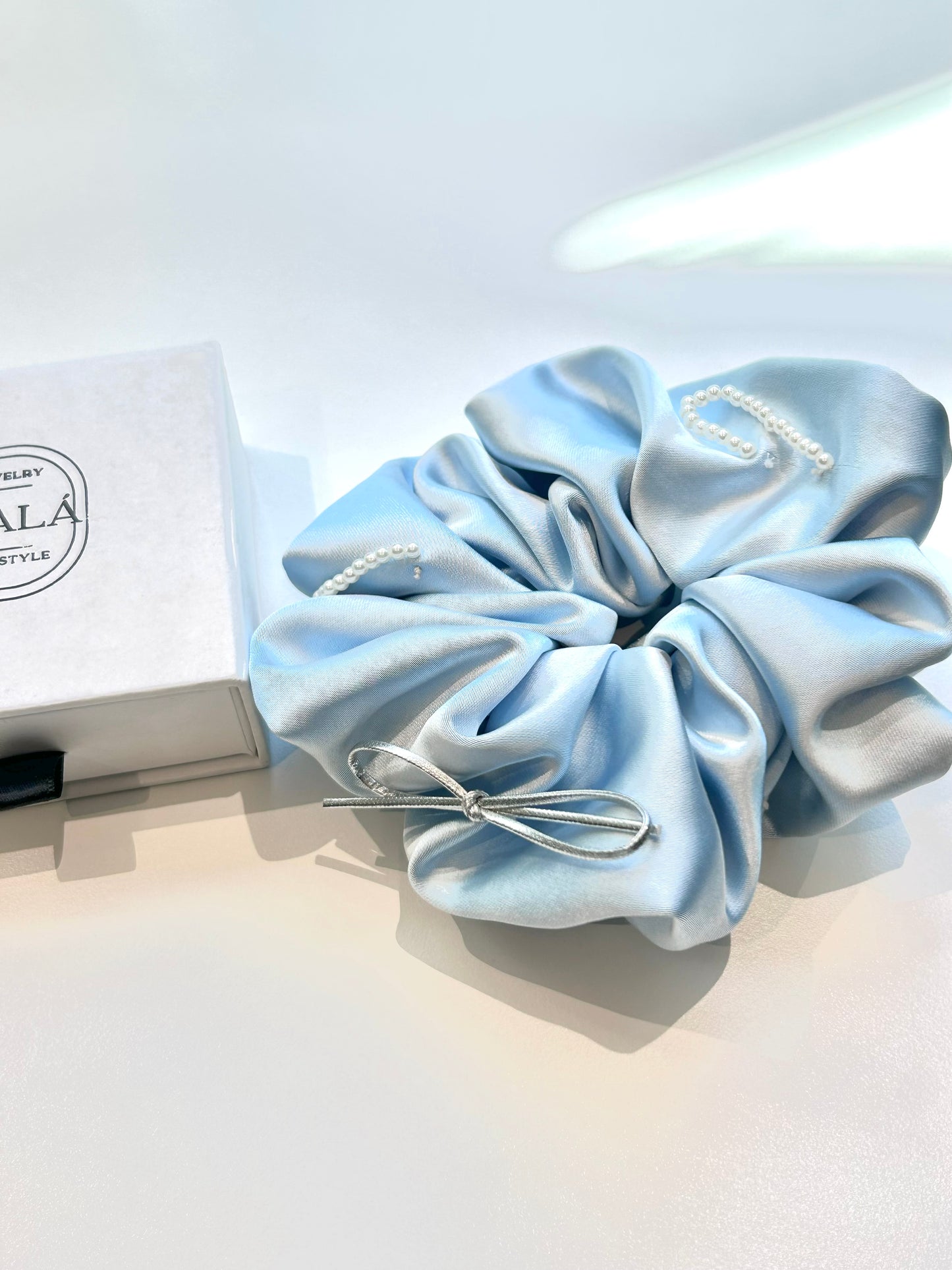A light blue satin hair scrunchie with pearl embellishments and a silver bow, shown next to its white branded gift box on a clean white surface.