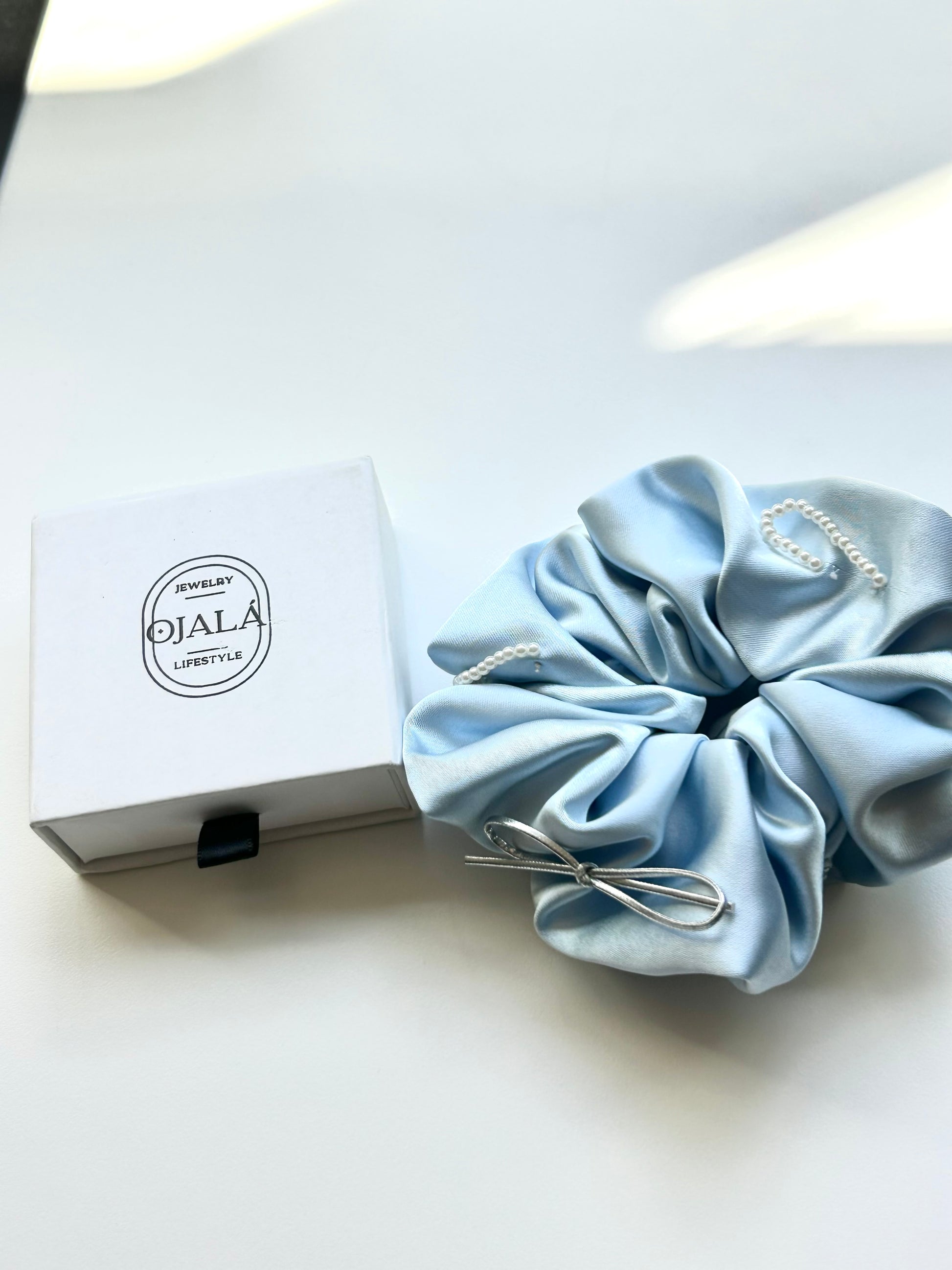 A light blue satin hair scrunchie with pearl-like bead accents and a silver metal bow, displayed next to its white branded box on a clean white surface.